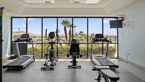 Ein Fitnessstudio mit Laufbändern, Crosstrainern und Bänken, mit großen Fenstern und Blick auf Palmen und einen Strand.