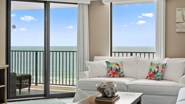 Ein Wohnzimmer mit einem weißen Sofa, bunten Kissen, einem kleinen Tisch mit einem Buch und einer dekorativen Schale, und Blick auf das Meer durch große Fenster.