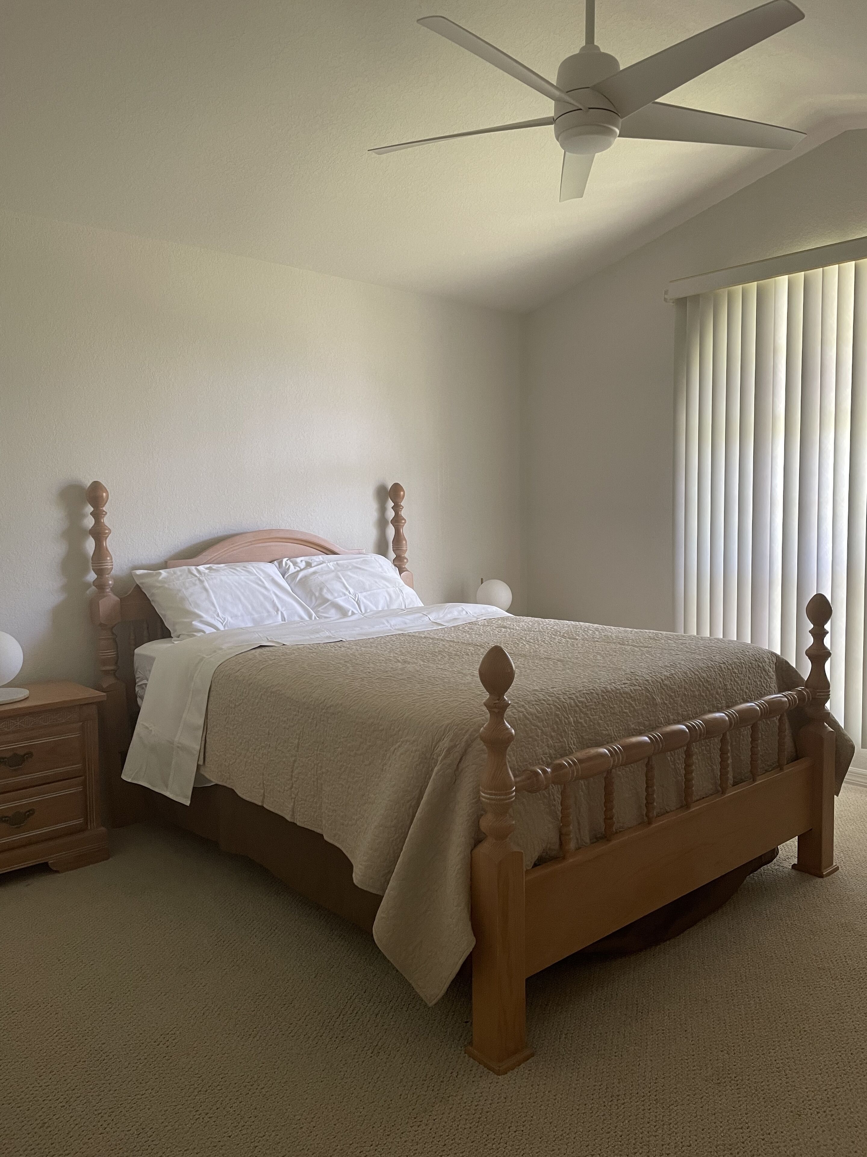 A bedroom with a wooden bed, white bedding, a nightstand, a ceiling fan, and a window with blinds.