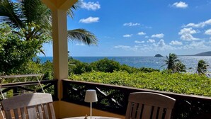 Mesa redonda de madera con sillas y una lámpara de mesa pequeña en un balcón con vistas a un paisaje tropical, palmeras y el océano.