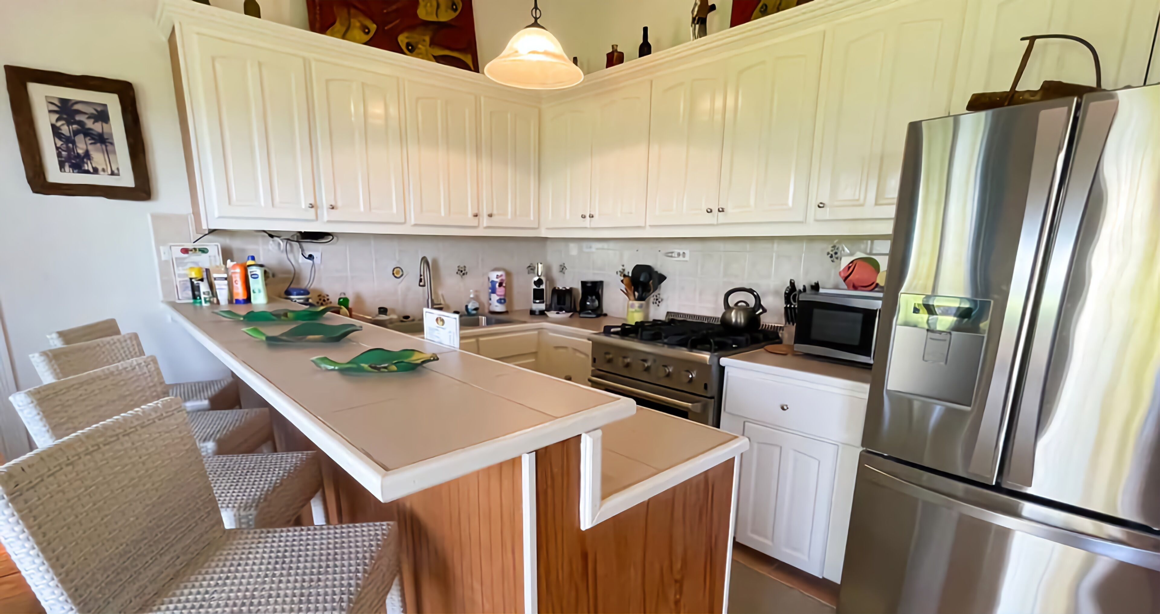 A kitchen with white cabinets, a stainless steel refrigerator, a stove, and a microwave. There are wicker chairs and a white countertop.