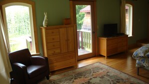 Une pièce avec une commode en bois, un téléviseur posé sur un meuble, une chaise et une grande fenêtre.