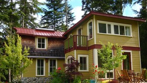 Une maison à deux étages, avec des fondations en pierre, un toit rouge et un jardin où poussent différentes plantes et fleurs.