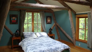 Une chambre avec un plafond en bois, un lit recouvert d’une couvre-lit à motifs floraux, deux tables de chevet et deux fenêtres avec des rideaux.