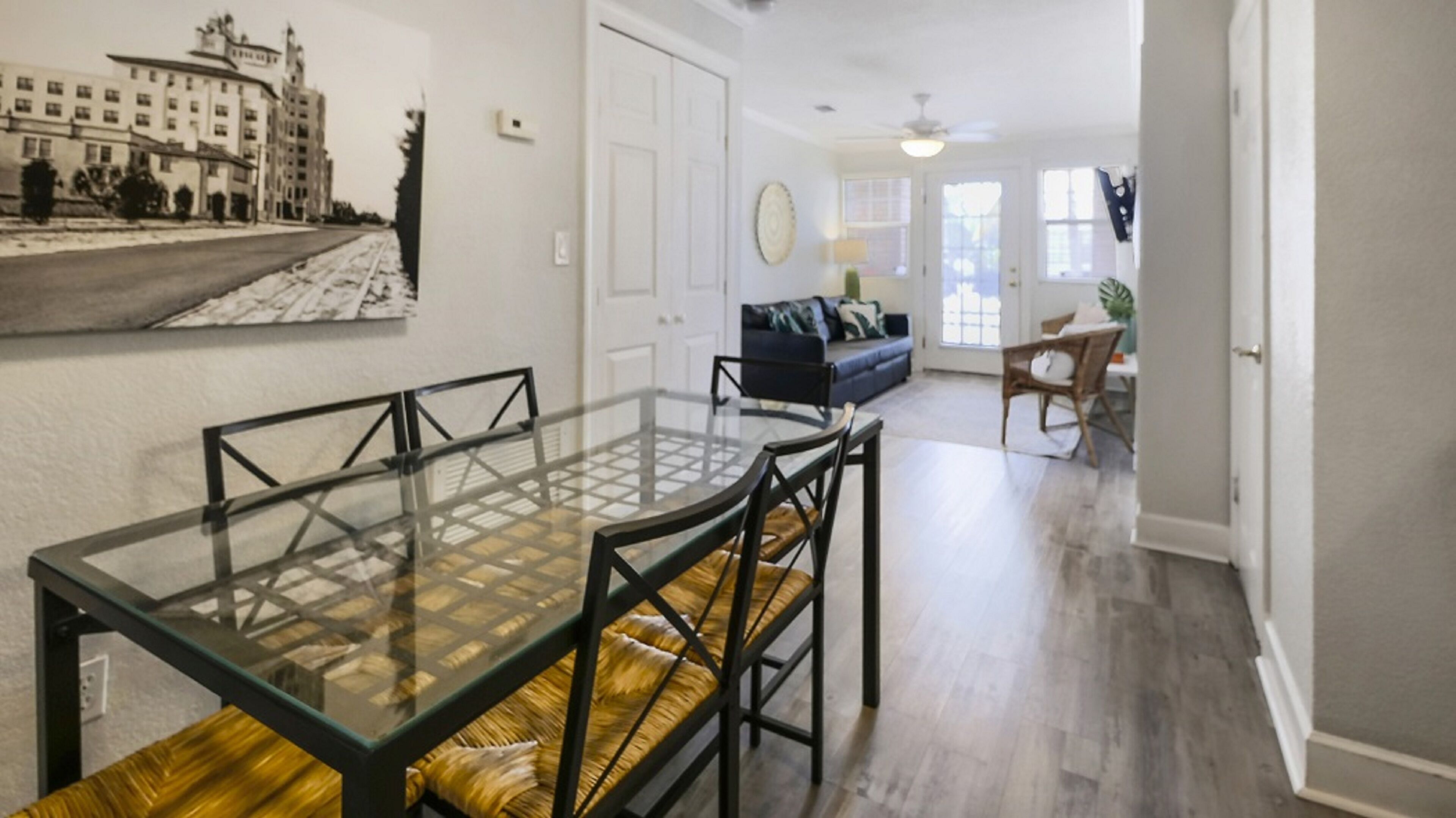 A dining area with a glass-top table and black chairs, a living room with a sofa and a chair, and a hallway leading to a door.