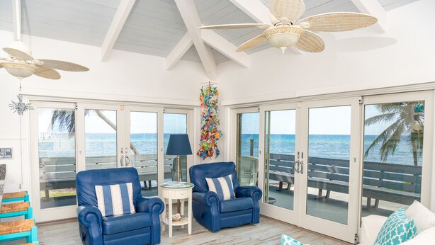 Amplia habitación con un gran ventanal corredera que da a un balcón con vistas al océano. Hay dos sillones azules y un sofá blanco con cojines de diseño azul y blanco.