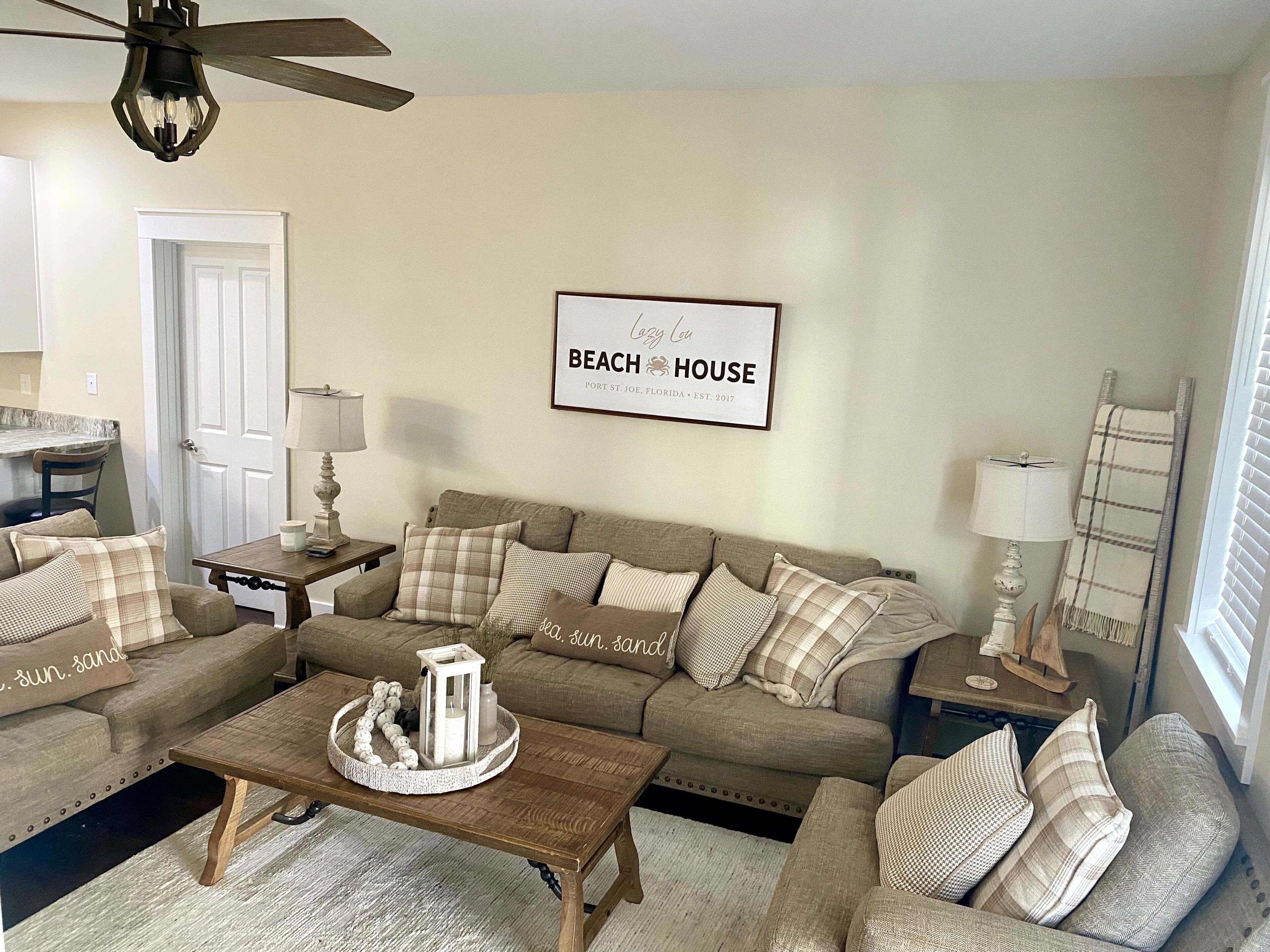 Uma sala de estar aconchegante com um sofá bege, uma mesa de centro de madeira e uma placa emoldurada com a inscrição 'Beach House'.