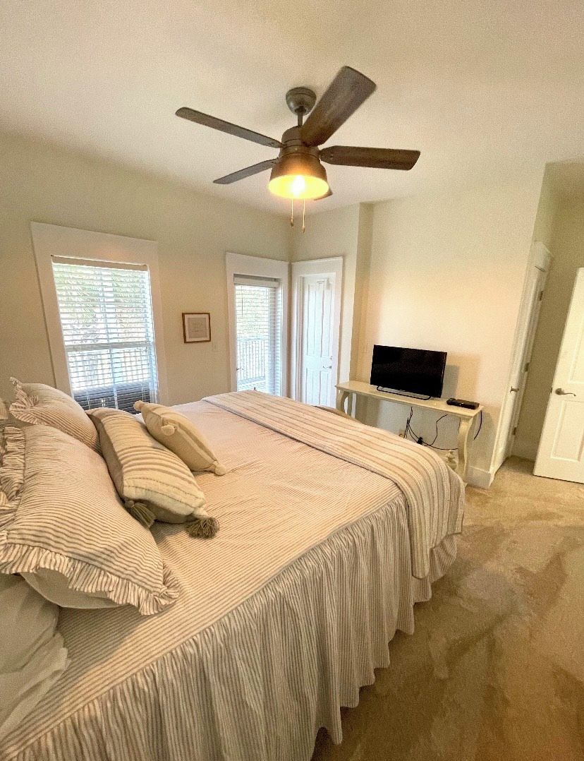 Une chambre à coucher avec un grand lit, un ventilateur de plafond, un téléviseur posé sur un meuble et une fenêtre avec des stores.