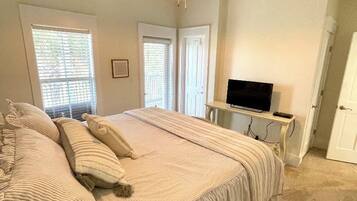 Une chambre à coucher avec un grand lit, un ventilateur de plafond, un téléviseur posé sur un meuble et une fenêtre avec des stores.
