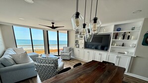 A modern living room with a large wooden dining table, a sofa, a patterned armchair, and a TV on a white cabinet. - Oceanfront Premium 3BR- Gourmet Kitchen! Farmhouse Decor! Gorgeous View! (Myrtle Beach)