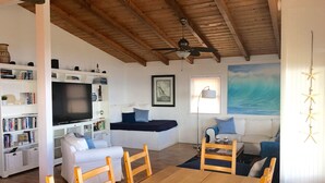 Un saló confortable amb sostre de fusta, un televisor de pantalla plana, un sofà, una taula de dinar amb caires i una estanteria plena de llibres.