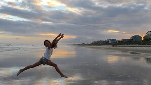 Escena de platja amb una persona estirant-se a la sorra, cases al fons i un cel ennuvolat.