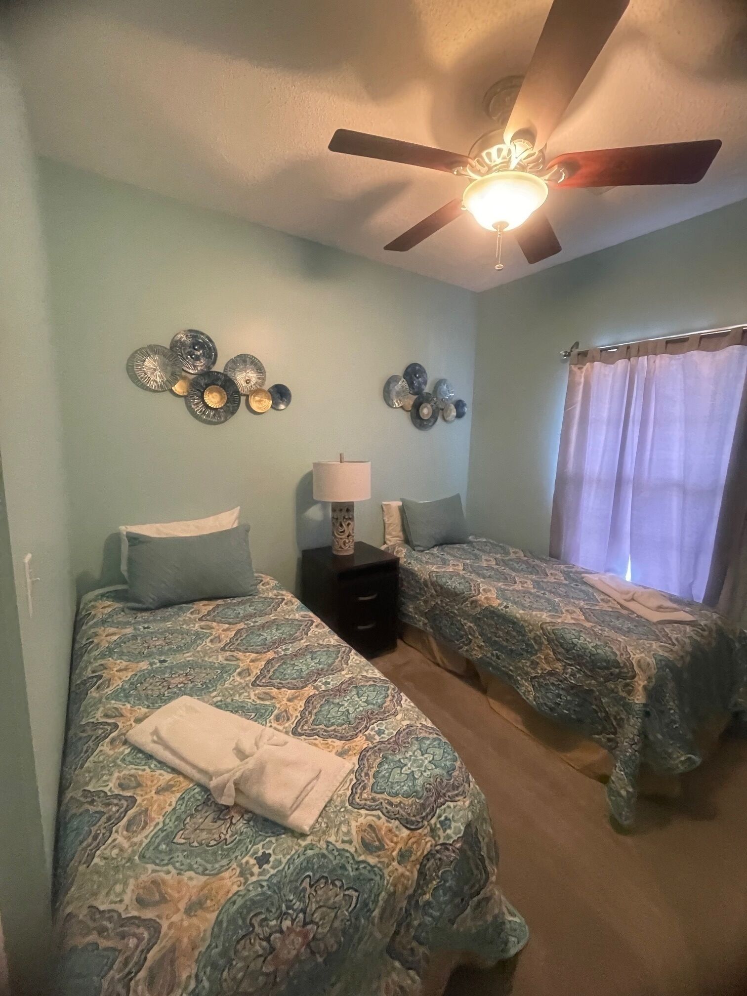 A bedroom with two beds, a ceiling fan, decorative wall art, and a window with curtains.