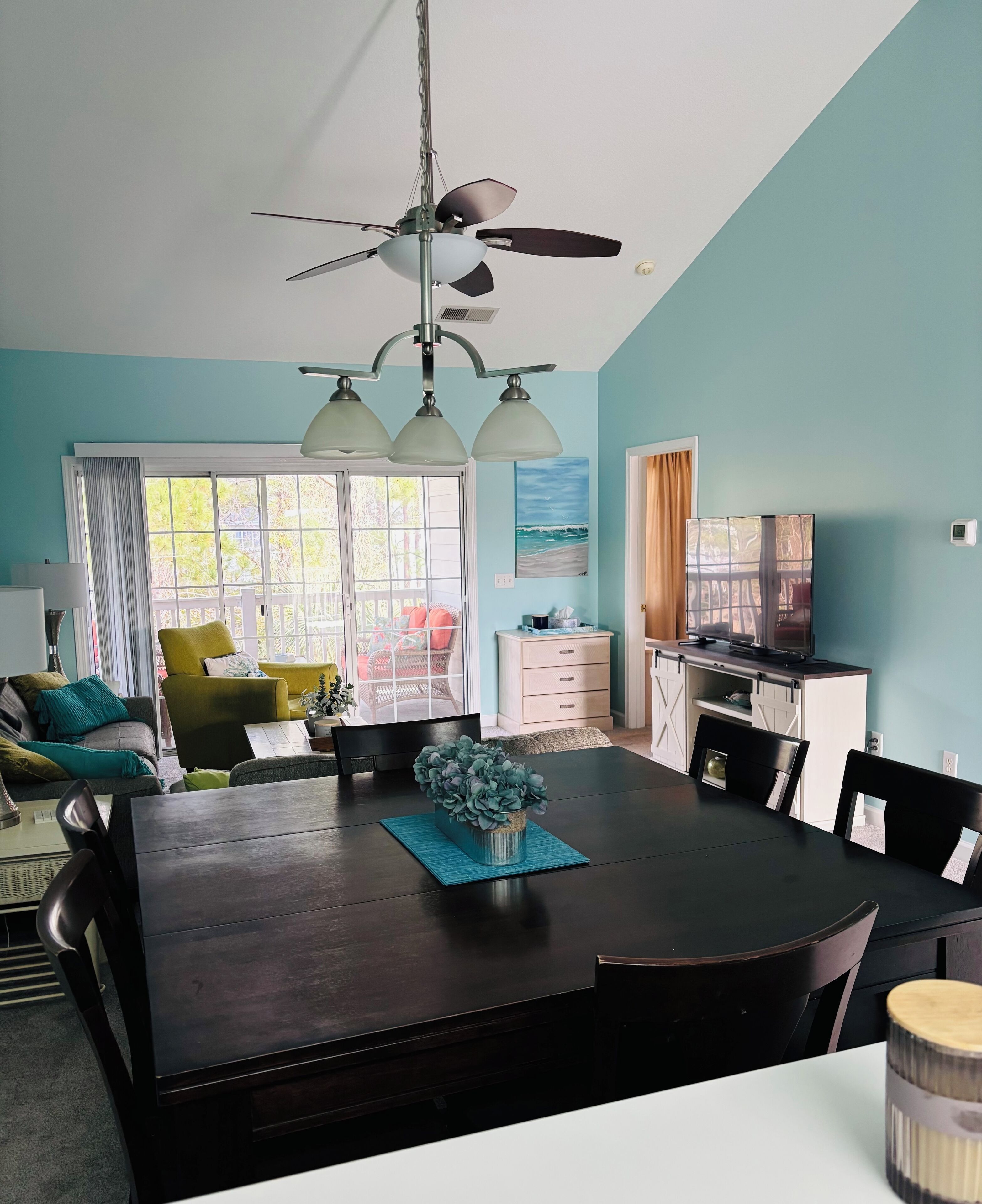A dining area with a dark wooden table, chairs, and a ceiling fan. There is a window with curtains and a view of the outdoors.