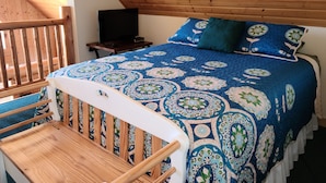 Quarto com teto inclinado, estrutura de cama de madeira, colcha de cama com padrão azul e branco e uma pequena mesa de cabeceira com uma televisão.