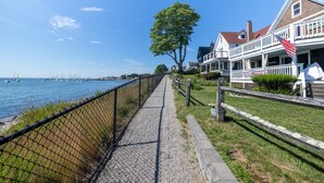 A waterfront walkway with a fence, a large tree, and houses with American flags.