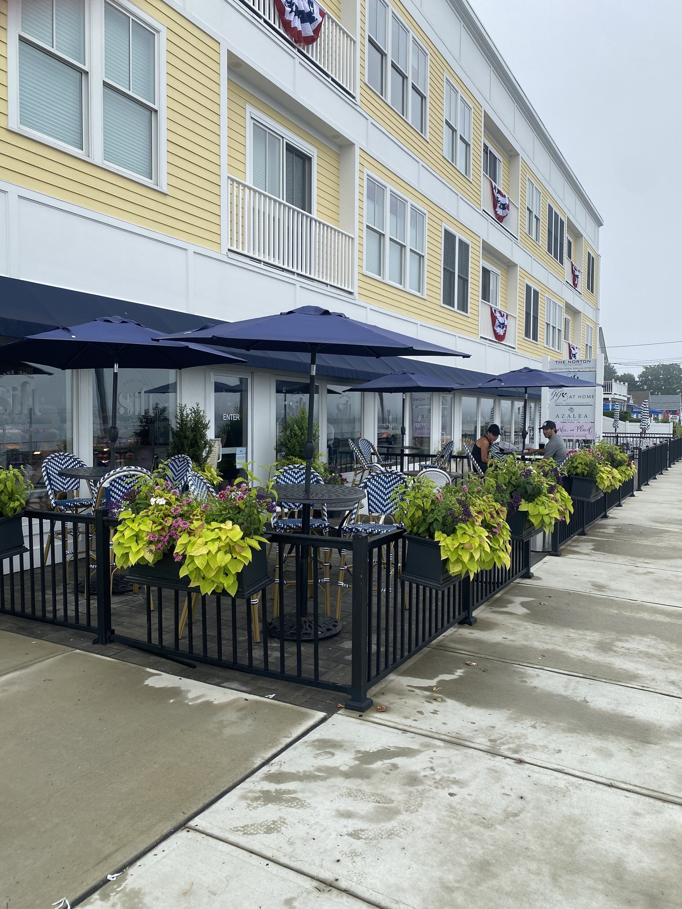 A row of outdoor tables with umbrellas, potted plants, and a building with yellow siding and white trim.