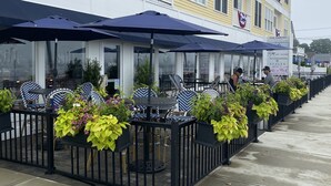 A row of outdoor tables with umbrellas, potted plants, and a building with yellow siding and white trim.