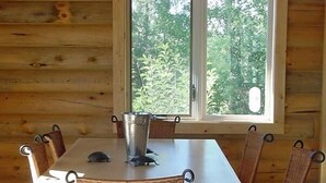 A wooden cabin interior with a dining table, wicker chairs, and a large window offering a view of greenery. - Secluded Lakefront Log Cabin, Wildlife and Fishing Paradise, Pet Friendly (Ashton)