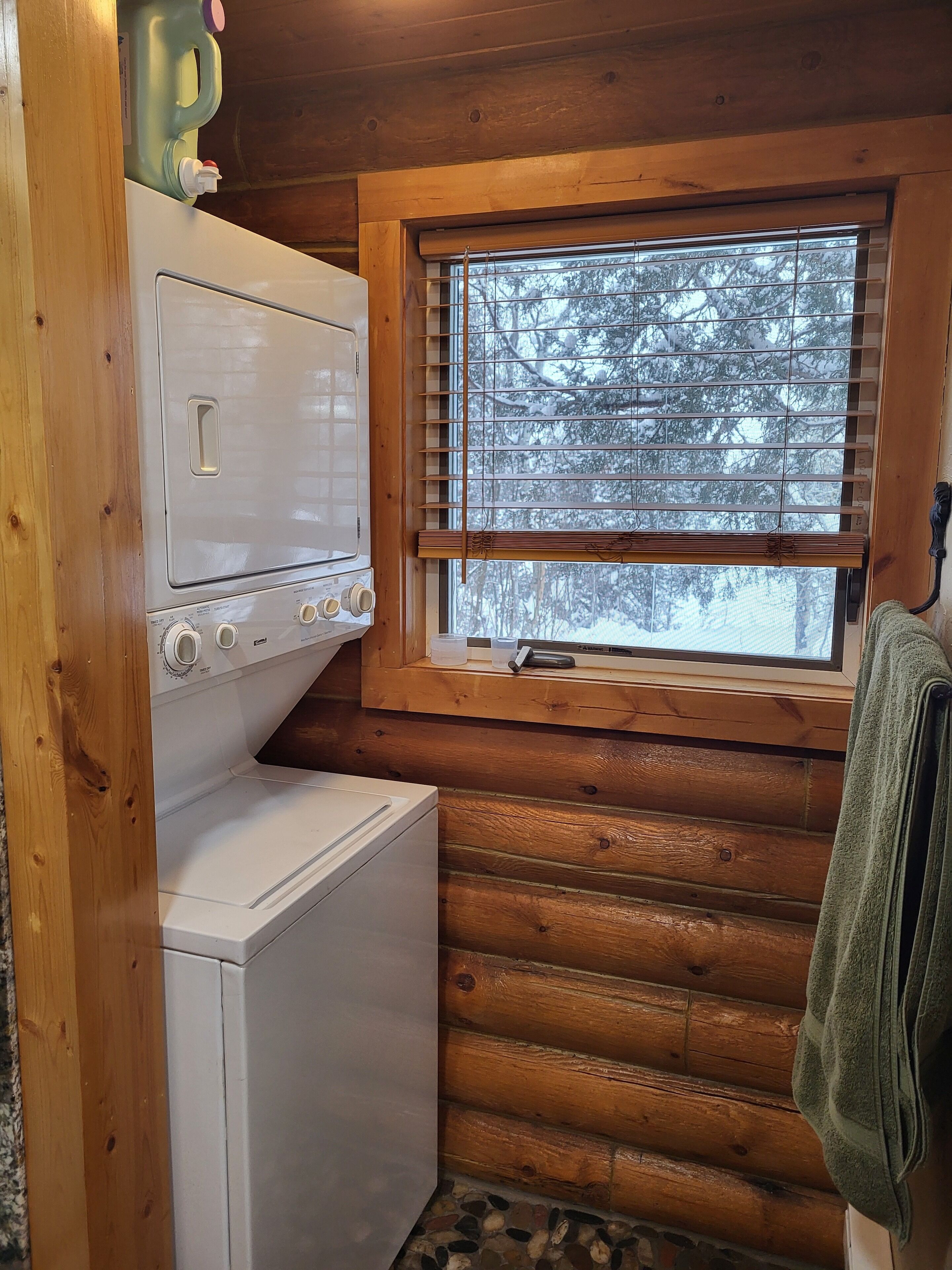A laundry room with a washer and dryer, wooden walls, a window with blinds, and a green towel hanging on the wall.