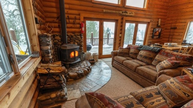 A log cabin interior with a stone fireplace, a wooden couch with patterned cushions, and a view of the outdoors through large windows. - Secluded Lakefront Log Cabin, Wildlife and Fishing Paradise, Pet Friendly (Ashton)