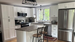 A modern kitchen with white cabinets, a stainless steel refrigerator, a large island with bar stools, and a window providing natural light. - "Moss Mountain Cottage" Clean, Private, Central MDI - 2025 updated kitchen (Bar Harbor)