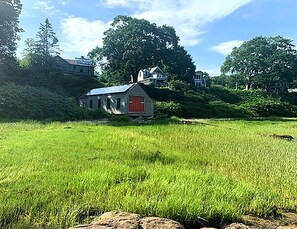 Una casa pequeña con puerta roja y techo inclinado, rodeada de campos verdes exuberantes y árboles.