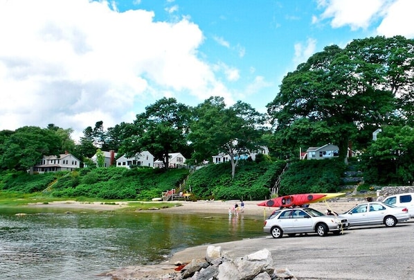 Cena à beira de um lago, com casas, uma praia de areia e carros estacionados.