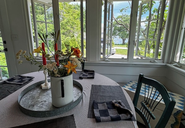A dining area with a table set for two, a vase with flowers, and a view of a lake and trees through large windows. - Ocean View Cottage Falmouth Foreside (Falmouth)