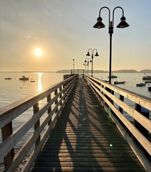 Un muelle de madera que se adentra en aguas tranquilas, con barcos y una costa lejana.