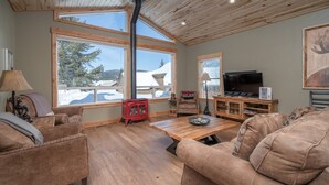 Una sala de estar acogedora con pisos de madera, ventanas grandes, chimenea y televisor.
