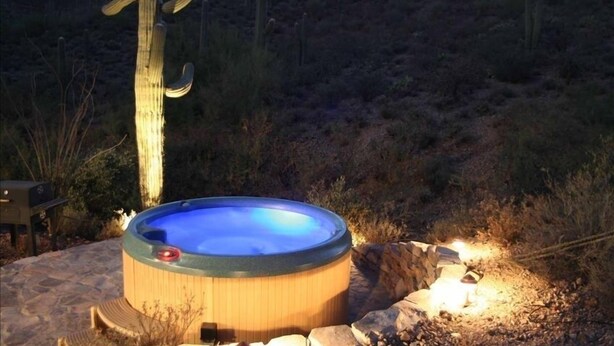 A hot tub in a desert setting with a wooden deck and a stone fire pit. - Sahuaro Ranch Hideaway (Tucson)