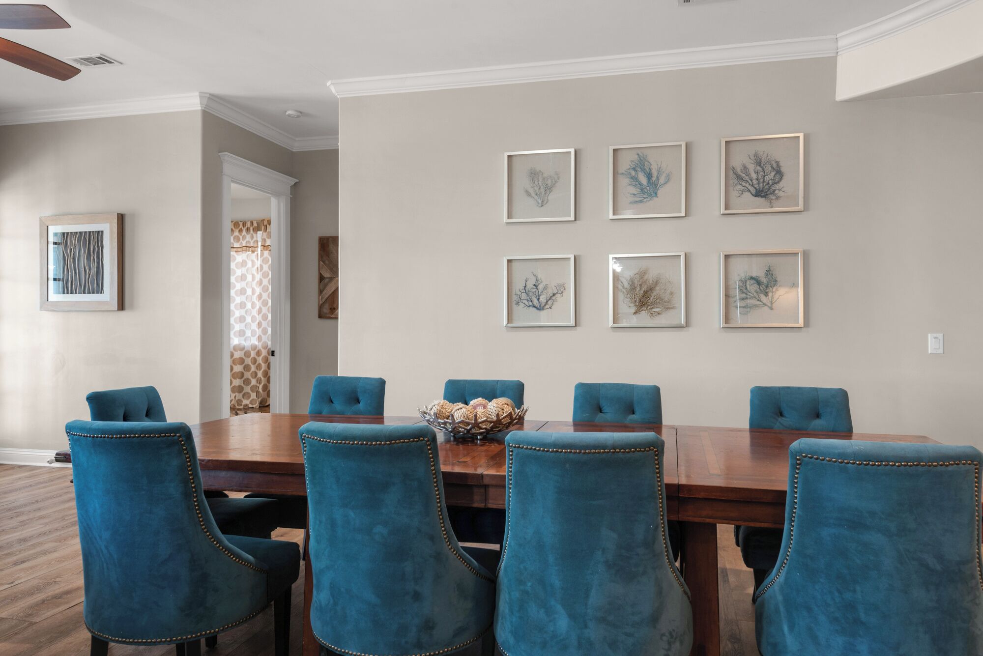 A dining room with a wooden table and six blue upholstered chairs. There are six framed artworks on the wall above the table.