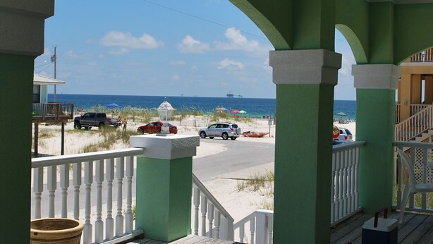 A view from a porch with green pillars and white railings, overlooking a beach with parked cars and umbrellas. - Oh the views, hear the surf Great location all year long (Gulf Shores)