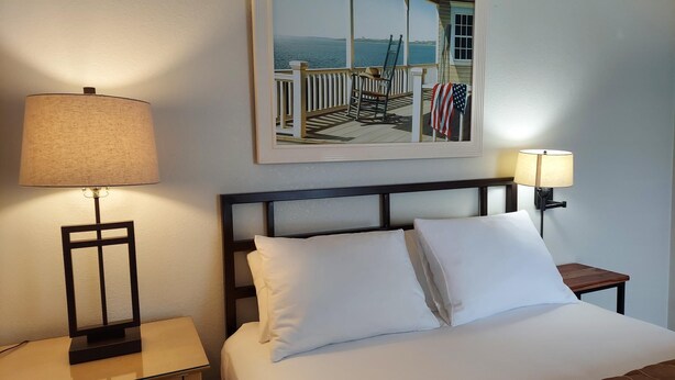 A neatly made bed with white linens, a framed painting of a coastal scene above it, and two bedside lamps on either side. - Luxury Townhouse at the Gateway to the Utah Canyons, Best Price On VRBO! (Cottonwood Heights)