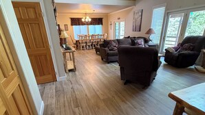 Amplia sala de estar con piso de madera, comedor con sillas y mesa, y un gran ventanal con persianas.