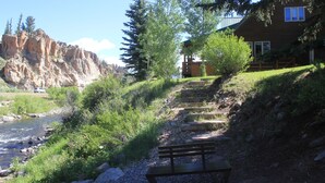 Un sendero junto al río con un banco de madera, una casa con terraza y un acantilado rocoso al fondo.