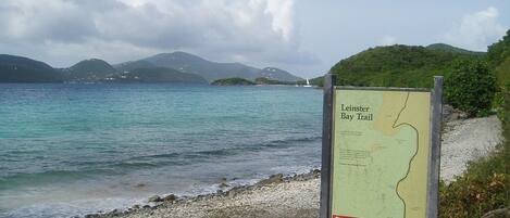 Un panneau indicateur intitulé « Leinster Bay Trail », avec une carte, est installé près d’une plage aux eaux cristallines, avec des collines qui se dessinent à l’horizon.
