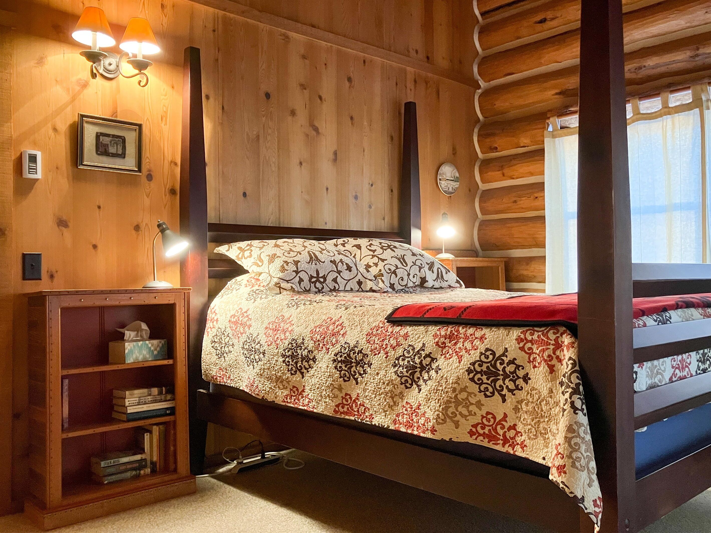 Een houten slaapkamer met een bed, nachtkastje, lamp, plank met boeken en een ingelijst schilderij aan de muur.