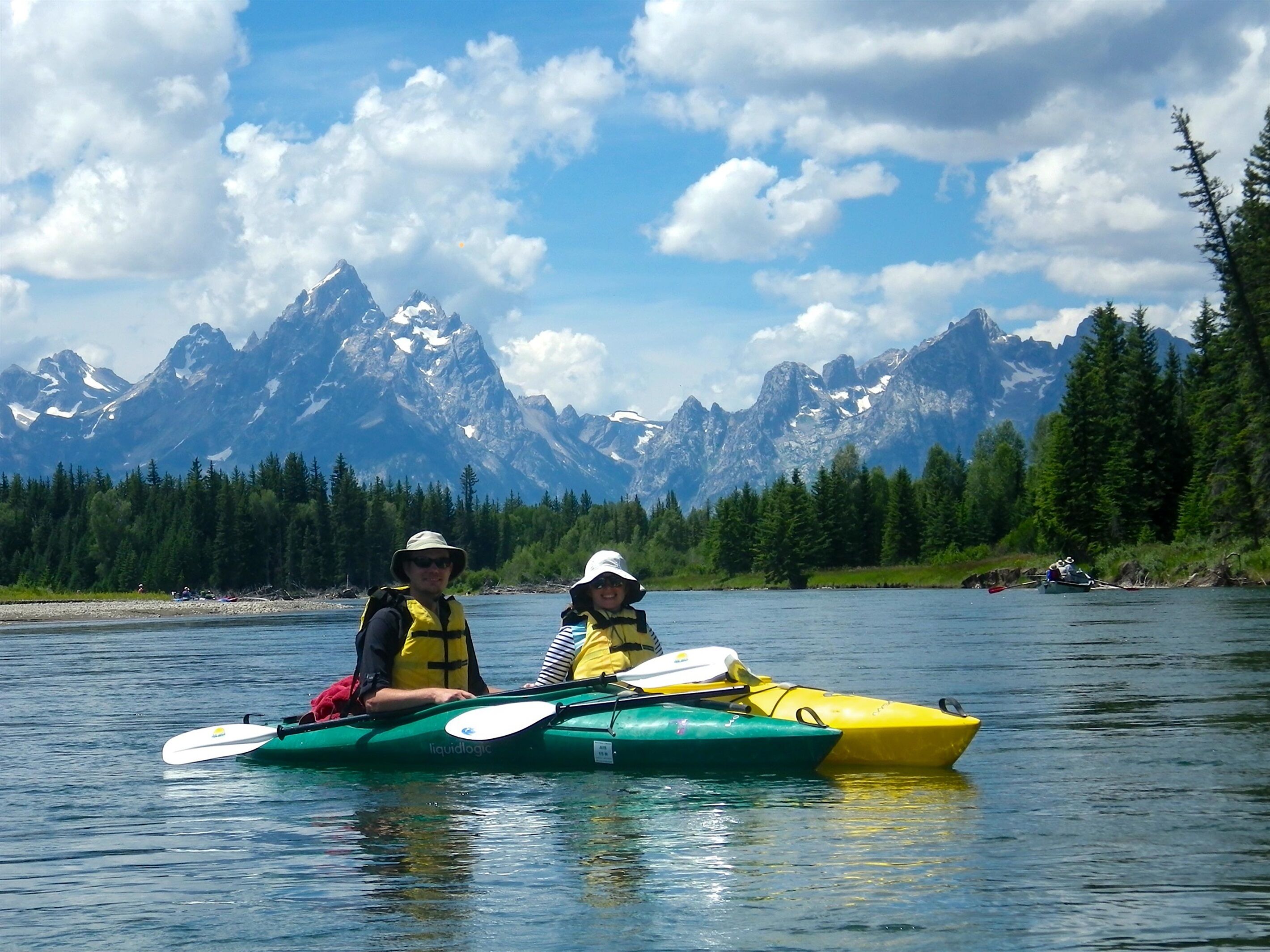 Two people kayaking on a calm river with a mountainous backdrop.