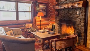 Ein gemütliches Blockhaus-Interieur mit Kamin aus Stein, Holzmöbeln und Blick ins Freie.