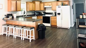 A modern kitchen with wooden cabinets, a central island with bar stools, a white refrigerator, and a black trash bin.