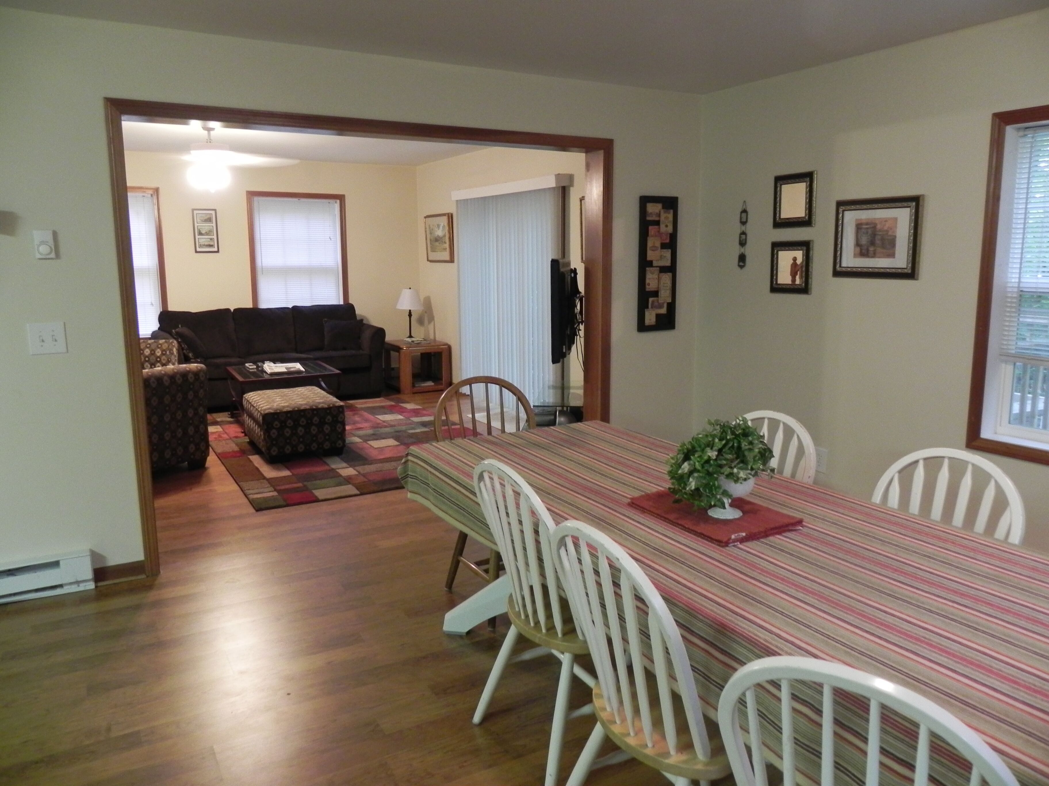A dining area with a striped tablecloth, white chairs, and a plant centerpiece. There is a living room in the background with a couch and a lamp.