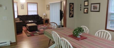 A dining area with a striped tablecloth, white chairs, and a plant centerpiece. There is a living room in the background with a couch and a lamp.