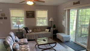 A living room with a sofa, two armchairs, a coffee table, and a ceiling fan. There are two windows with blinds, a lamp, and a welcome mat near the door.