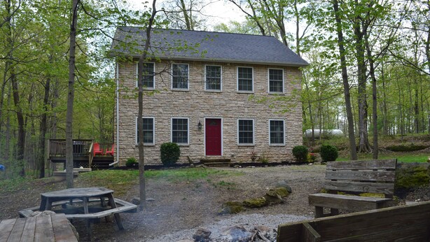 A stone building with a red door, surrounded by trees and a fire pit area with benches.