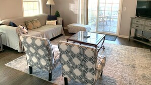 A living room with a sofa, two patterned armchairs, a coffee table, a TV, and a sliding glass door leading to a patio.