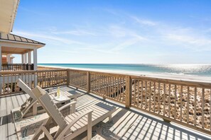 Terraza de madera con sillones de descanso y una pequeña mesa, con vista a una playa de arena y aguas azules cristalinas.