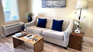 Ett vardagsrum med en beige soffa, två blå kuddar, ett träbord, en golvlampa, en krukväxt och en tavla på väggen.