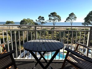 A balcony with a wooden table and chairs, overlooking a pool and a beach with trees. - Spectacular Oceanfront, Top Floor, 1 BR, 1 BA, steps from the beach! (Hilton Head Island)